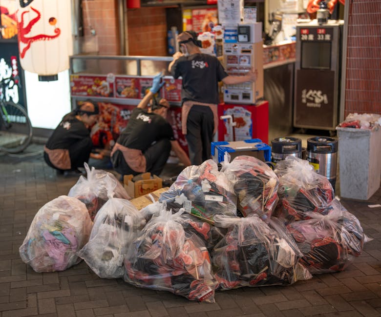 A close-up view of a roadside rubbish pile consisting of black plastic garbage bags, broken pieces of cardboard, and scattered debris on a dark asphalt surface. A yellow plastic storage container and