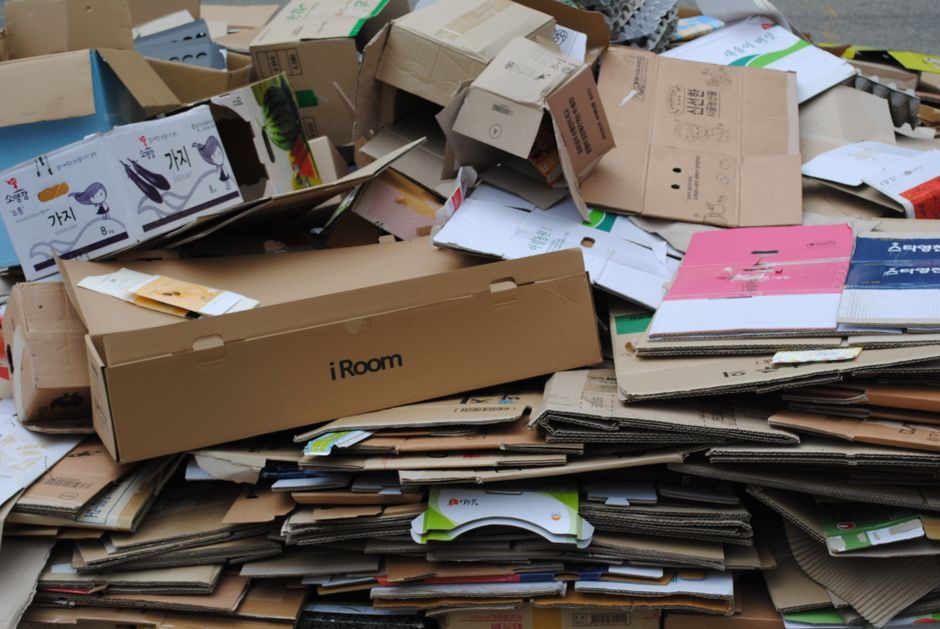 A large pile of flattened cardboard boxes and packaging materials is stacked outdoors on a paved surface. The cardboard varies in size, color, and finish, with some boxes displaying printed branding a