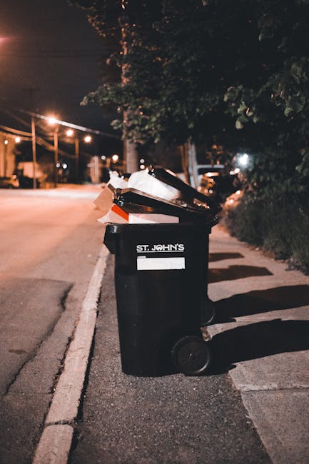A large pile of flattened cardboard boxes and packaging materials is stacked outdoors on a paved surface. The cardboard varies in size, color, and finish, with some boxes displaying printed branding a